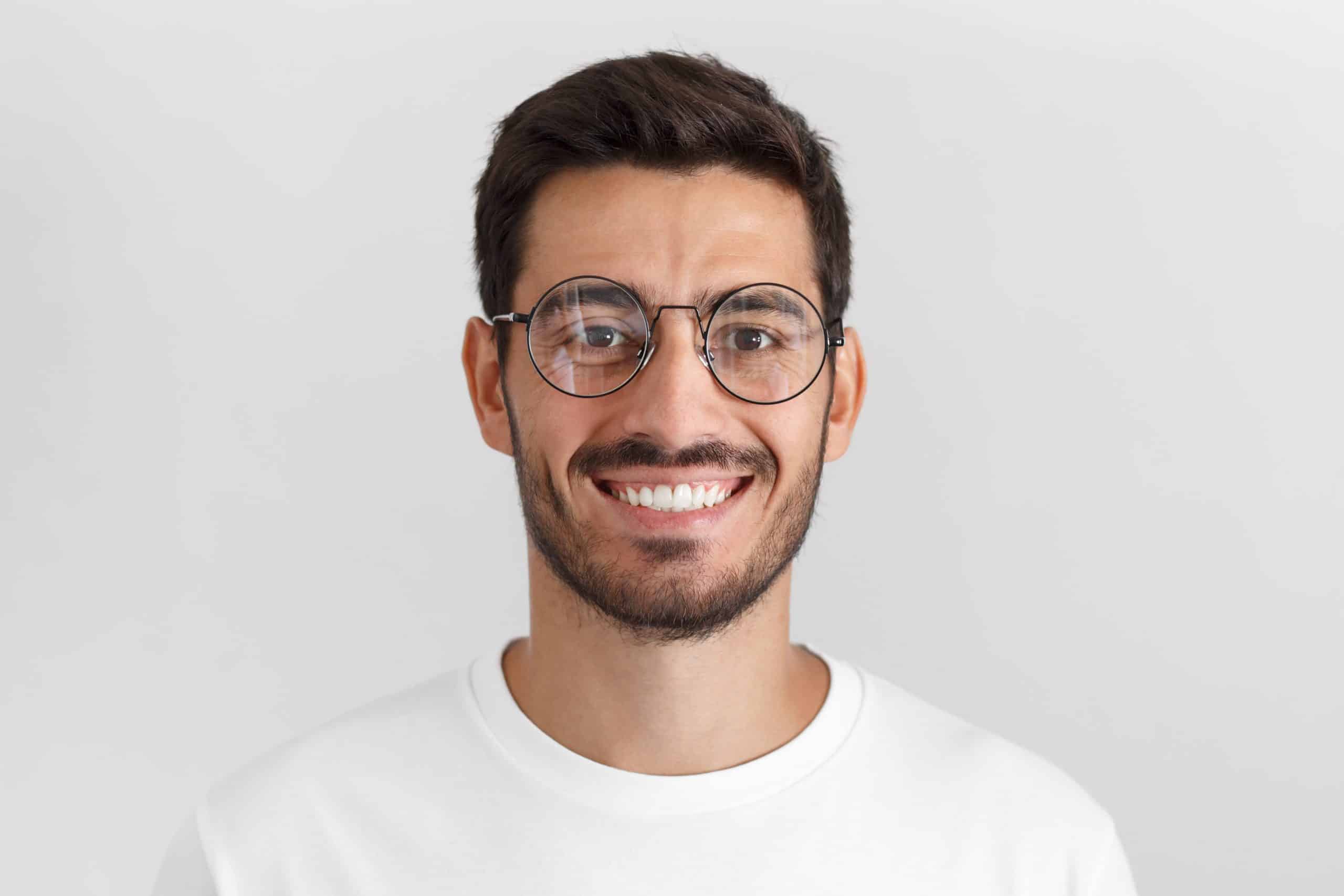 Daylight portrait of young handsome caucasian man isolated on grey background, dressed in white t-shirt and round eyeglasses, looking at camera and smiling positively